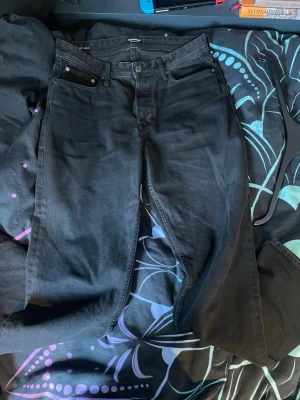Svarta jeans från Jack and Jones  - Säljer ett par svarta raka jeans från Jack and Jones . Klassisk femficksmodell med knappgylf och diskret logga på bakfickan. Jeansen har en clean look och är tillverkade i kraftig denim. Perfekta för dig som gillar minimalistisk stil. Perfekt Jens till en stilrent outfit. Priset är inte huggit i sten och kan diskuteras.