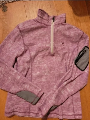 Lila fleece-tröja med halv dragkedja - Mysig lila fleece-tröja med halv dragkedja framtill och hög krage. Tröjan har en praktisk ficka med dragkedja på ena ärmen och gråa detaljer vid ärmslut och ficka. Perfekt för kyliga dagar och outdoor-aktiviteter.