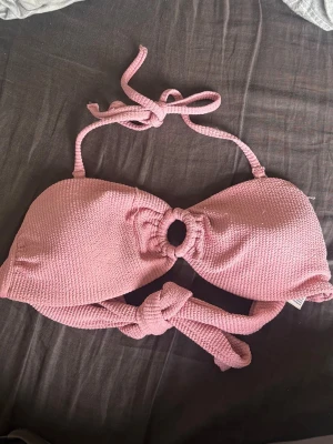Rosa ribbad bikiniöverdel med knyt - Säljer en snygg rosa bikiniöverdel med ribbad struktur och knyt både i nacken och framtill. Toppen har en rund ringdetalj i mitten som ger en trendig look. Perfekt för lata dagar på stranden eller poolen. Den har skavanker (se bilder), därav priset 