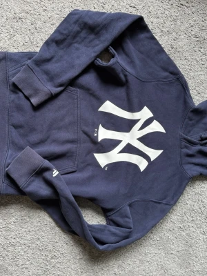 Mörkblå New York Yankees hoodie - Mörkblå hoodie med stor vit New York Yankees-logga på bröstet. Klassisk känguruficka framtill och ribbade muddar vid ärmar och nederkant. Tillverkad i mjukt sweatshirtmaterial och har en huva för extra komfort.