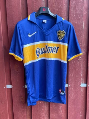 Blå Boca Juniors matchtröja Nike - Snygg Boca Juniors matchtröja i blått med bred gul rand och vita detaljer samt stor krage. Tröjan har klubbmärke på bröstet, Nike-logga och tryckta sponsorloggor 'Quilmes' och 'Xeneizes'. Storlek L
