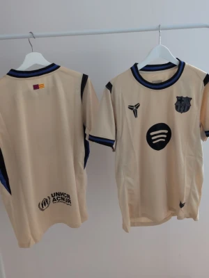 Beige FC Barcelona matchtröja - Helt ny Snygg beige FC Barcelona matchtröja från Nike med svarta och blå detaljer. Tröjan har klubbmärke, Spotify-logga och UNHCR-tryck samt korta ärmar och rund hals. Perfekt för dig som älskar fotboll och vill ha en unik supportertröja.