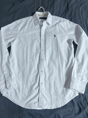 Vit skjorta från Ralph Lauren - Klassisk vit skjorta från Ralph Lauren med broderad logga på bröstet. Skjortan har lång ärm, knappar framtill och en tidlös passform. Perfekt till jeans eller chinos för en clean och stilren look.
