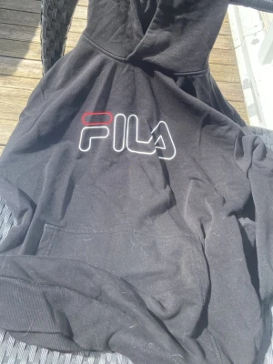 Svart hoodie från FILA med logga - Säljer en svart hoodie från FILA med stor vit och röd logga på bröstet. Hoodien har klassisk huva och magficka framtill. Perfekt för dig som gillar streetwear och vill ha en enkel men snygg look.
