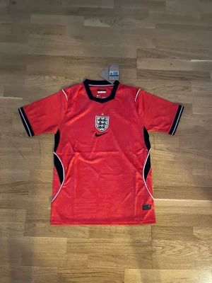 Röd England Nike fotbollströja - Snygg röd England fotbollströja från Nike med svart och vit detaljering. Tröjan har korta ärmar, svart krage och England-emblemet på bröstet. Tillverkad i lätt och ventilerande material, perfekt för träning eller match.        Fraktar dagen efter eller samma dag som du köper. 