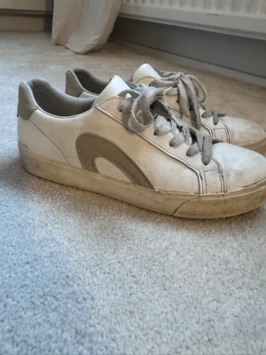Vita sneakers med grå/beiga detaljer - Snygga vita sneakers med grå detaljer. Skorna har en låg profil och en klassisk siluett som passar till det mesta. Tillverkade i läder och textil för en bekväm känsla hela dagen. Tvättat dom någon gång men grov tvätt får göras själv!!