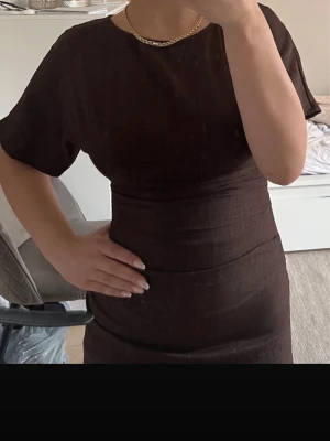 Brun långklänning med kort ärm - Snygg brun långklänning med korta ärmar och rund halsringning. Klänningen har en figurnära passform och är tillverkad i ett mjukt tyg som ger en elegant siluett. Perfekt för dig som gillar stilrena och enkla plagg.