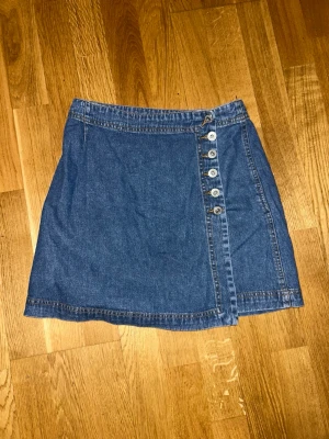Jeanskjol med knappar från Hennes - Snygg blå jeanskjol från Hennes collection i omlottmodell med fem knappar framtill. Kjolen är kort och har klassisk denimlook, perfekt att matcha med en t-shirt eller hoodie. Gjord i bomull för en skön känsla.