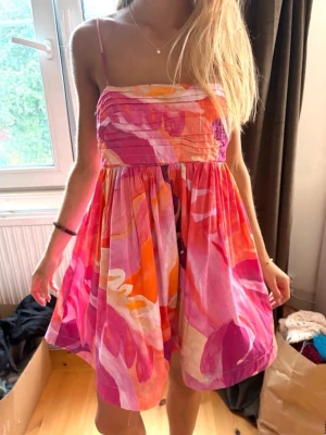 Färgglad kort sommarklänning - Säljer en superfin kort klänning med tunna axelband och färgstarkt mönster i rosa, orange, rött och vitt. Klänningen har en luftig passform och är perfekt för varma dagar. Materialet känns lätt och bekvämt mot huden.