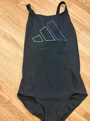 Svart baddräkt från Adidas - Snygg svart baddräkt från Adidas med tunna axelband och stort logotryck i neonfärger på bröstet. Modellen har en sportig rygg med öppen detalj och är tillverkad i ett stretchigt, snabbtorkande material som passar perfekt för simning och träning.