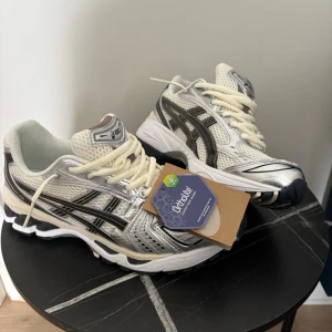 Asics gel kayano 14 cream mettalic plum (ingen box) - Snygga sneakers från Asics i vitt och silver med svarta detaljer. Skorna har en sportig design med mesh- och syntetmaterial, samt klassiska Asics-stripes på sidorna. Tjock sula för extra komfort och snörning framtill. Perfekta för en trendig streetwear-look. ‼️⚠️Strl 41,5. Box kommer inte med⚠️‼️ jag använde dom 1 gång utom men tyckte ej om dom för dom inte passa min vardagsliv.