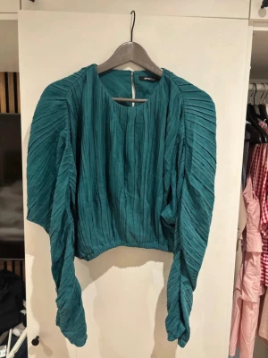 Grön plisserad croppad topp - Säljer en unik grön croppad topp med plisserade detaljer och långa, vida ärmar. Toppen har rund halsringning och ett coolt strukturerat tyg som ger en trendig look. Perfekt för dig som vill sticka ut med en statement piece.