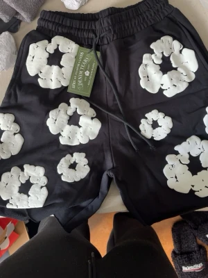 Denim tears shorts black  - Svarta shorts från Denim Tears med stora vita blommor i ett broderat mönster. Shortsen har elastisk midja med snörning för skön passform och är perfekta för en avslappnad stil.