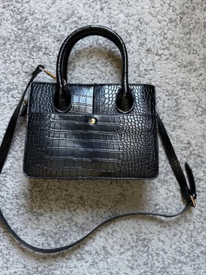Svart croco-mönstrad handväska - Stilren svart handväska med croco-mönster och både handtag och axelrem. Väskan har en fyrkantig form och en liten guldfärgad knappdetalj framtill. Perfekt för dig som gillar trendiga accessoarer med lyxig känsla.