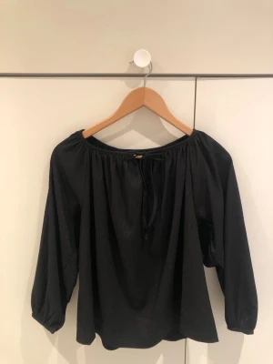 Svart offshoulder blus med knytband - Stilren svart offshoulder blus med långa ärmar och knytband framtill. Blusen har en lös passform och är tillverkad i ett lätt, mjukt tyg som ger en avslappnad look. Perfekt att matcha med jeans eller kjol för en trendig outfit.