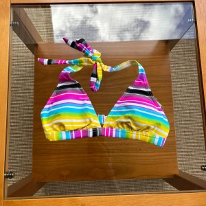 Randig bikiniöverdel från Race Marine - Bkiniöverdel från Race Marine med ränder i rosa, lila, gul, grön, blå, svart och vit. Triangelformade kupor, knyt i nacken och spänne i ryggen. Storlek 34. Oanvänd med prislapp.. Storlek 34.