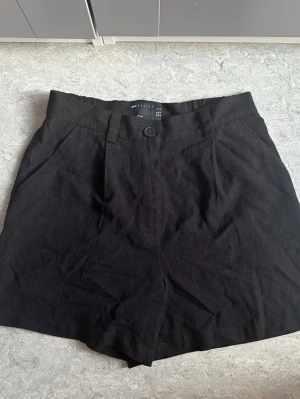 Svarta shorts  - Stilrena svarta shorts från ASOS DESIGN och knappstängning framtill. Klassisk rak passform med bälteshällor och sidofickor. Perfekta till sommaren och lätta att matcha med olika toppar. Den är petite så den passar för de som är korta. Oanvänd.