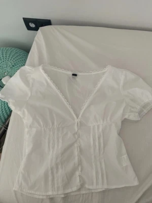Vit blus med puffärm och spetskant - Söt vit blus med puffärmar, knappar framtill och dekorativ spetskant längs urringningen. Blusen har figurnära passform och fina veckdetaljer framtill. Perfekt för en romantisk och trendig look.