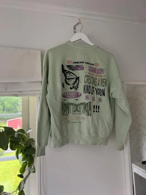 Mintgrön sweatshirt med tryck - Cool mintgrön sweatshirt med unikt grafiskt tryck på ryggen i flera färger, bland annat svart, lila och grönt. Tröjan har rund hals, ribbade muddar och avslappnad passform. Perfekt för dig som gillar streetwear och statement-plagg. Tröjan har några fläckar, se bilder 