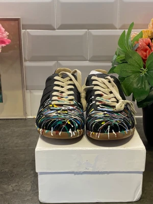 Maison Margiela sneakers med färgstänk - Unika svarta sneakers från Maison Margiela med färgstänk i vitt, blått, gult och grönt. Skorna har beige snörning och klassisk låg siluett. Tillverkade i läder med beige innersula och gummisula. Perfekta för dig som vill sticka ut med din stil.