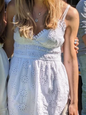 Vit sommarklänning med spetsdetaljer - Söt vit klänning med broderade spetsdetaljer och omlott-v-ringning. Klänningen har smala axelband och markerad midja med smock. Perfekt för soliga dagar och studentfirande.