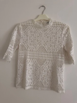 Vit spetsblus med trekvartsärm - en superfin vit spetsblus med trekvartsärm och rund hals. Blusen har ett detaljerat spetsmönster och knäppning i nacken. Perfekt för dig som gillar romantisk och elegant stil.