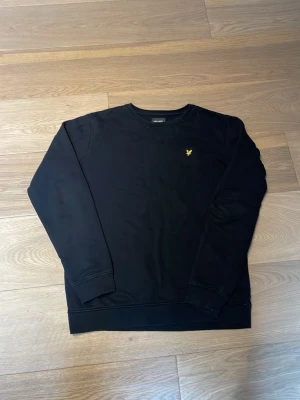 Svart sweatshirt från Lyle & Scott - Svart sweatshirt från Lyle & Scott med rund halsringning och klassisk gul logga broderad på bröstet. Tröjan har ribbade muddar vid ärmslut och nederkant. Perfekt för en stilren och avslappnad look. Sitter ungifär som en Smal 