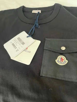 Svart Moncler t-shirt med ficka - Svart t-shirt från Moncler med klassisk rund halsringning och en snygg bröstficka med knapp och Moncler-logga. T-shirten är i bomull och har en stilren design som passar perfekt till jeans eller shorts. Helt ny aldig använd! Kom privat för frågor.