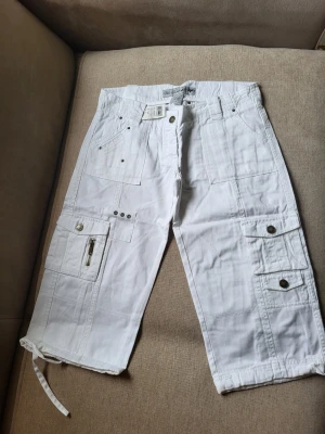 Vita cargoshorts med snörning Wet Seal size 3 US S - Säljer ett par vita cargoshorts med flera fickor, dragkedja och knappstängning. Shortsen har snörning vid bensluten och coola detaljer som nitar och ficklock. Perfekta för sommaren och riktigt sköna att bära.