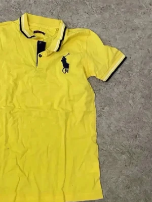 Gul piké från Polo Ralph Lauren - Snygg gul pikétröja från Polo Ralph Lauren med klassisk krage och knappslå. Tröjan har korta ärmar och den ikoniska polospelaren broderad i marinblått på bröstet. Perfekt för en avslappnad men stilren look.