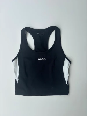 Svart sport-bh från Björn Borg - Snygg svart sport-bh från Björn Borg med vita paneler på sidorna och racerback-design. Tillverkad i stretchigt funktionsmaterial som andas, perfekt för träning och sportaktiviteter. Logga framtill i vitt.