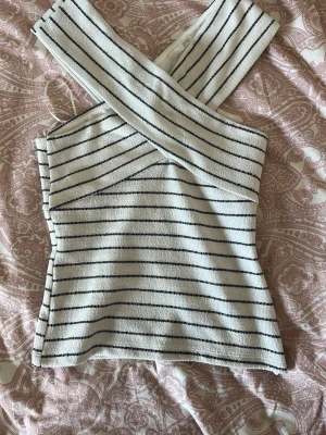 Randig omlottopp från Zara - Säljer en snygg vit omlottopp från Zara med svarta horisontella ränder. Toppen har en unik omlottdesign över axlarna och är ärmlös. Säljer för att jag köpte en för liten storlek och tog redan bort lappen innan.