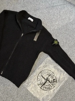 Stickad ziptröja från Stone Island - Säljer en ribbstickad ziptröja från Stone Island med ikonisk logotyp på ärmen. Finns i både ljusgrått och svart. Tröjan har hög krage, hel dragkedja och är tillverkad i mjukt stickat material. Perfekt för lager-på-lager och streetwear. Har färgen grå och svart. Pris 1800 för båda storlek s men funkar till M. Jag är 170 och de passar mig så de passar från 168 till 175. Pris kan disktueras.