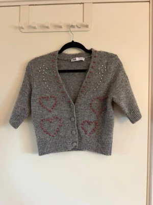 Grå kofta med pärlhjärtan från Zara - Kort grå kofta från Zara med v-ringning och trekvartsärmar. Dekorerad med pärlor i form av hjärtan och prickar i rosa och vitt framtill. Stängs med fyra knappar och har ribbade muddar. Perfekt för dig som gillar gulliga detaljer och en mysig look. Använd en gång. Storlek S men sitter som XS