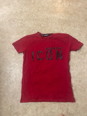 Röd ICON t-shirt från Dsquared2 - Snygg röd t-shirt från Dsquared2 med svart text 'ICON' och 'IBRAHIMOVIC' på bröstet. På ryggen finns ett stort svartvitt tryck av en person och texten 'DSQUARED2' upptill. Klassisk passform och korta ärmar. Perfekt för dig som gillar streetwear och statement-plagg.