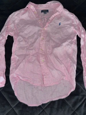 Rosa linneskjorta från Polo Ralph Lauren - Klassisk rosa skjorta från Polo Ralph Lauren i linne med blå broderad logga på bröstet. Skjortan har knappar framtill och lång ärm, perfekt för en avslappnad men ändå stilren look. Passar till killar 10-12 år som använder M. 