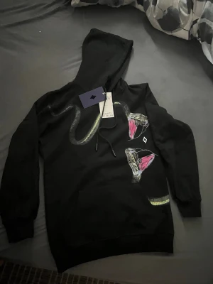 Marcelo Burlon hoodie - Detta är en Marcelo burlon hoodie helt ny, aldrig använd med taggarna och allt hängandes kvar. Den är svart med ormdetaljer på väldigt unik och fräsch.