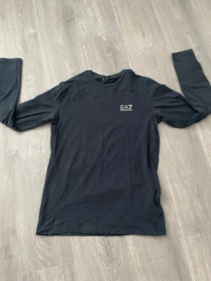 Svart långärmad topp från EA7 - Snygg svart långärmad topp från EA7 Emporio Armani med rund halsringning och diskret logga på bröstet. Tillverkad i mjukt material som känns skönt mot huden. Enkla vita ränder på ryggen nedtill ger en sportig touch.
