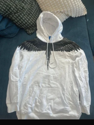 Marcelo burlon hoodie vit - Snygg vit hoodie från Marcelo Burlon med svart vingmotiv över axlar och bröst, samt på ryggen. Hoodien har huva med dragsko och en stor magficka. Perfekt för dig som vill sticka ut med en unik design. L passar M också 