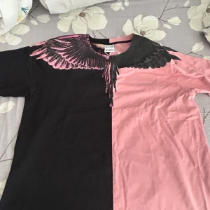 Svart och rosa t-shirt Marcelo Burlon - Unik t-shirt från Marcelo Burlon County of Milan med coolt fågelvinge-tryck över axlarna. Halva t-shirten är svart och andra halvan är rosa, vilket ger en riktigt edgy look. Rund hals och korta ärmar, tillverkad i mjuk bomull. Perfekt för dig som gillar statement-plagg.stolrke L/XL