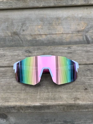Pastell Sportiga regnbågssolglasögon - Säljer ett par sportiga solglasögon med spegelglas i regnbågsfärger och vit båge. De har en futuristisk, kantig form och passar perfekt för dig som vill sticka ut. Linsen har en cool spegeleffekt och bågarna är i lätt syntetmaterial.
