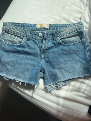 Blå jeansshorts från Miss Sixty - Säljer ett par klassiska blå, lågmidjade jeansshorts från Miss Sixty med råa, fransiga kanter och femficksmodell. Shortsen har normal passform och är tillverkade i slitstark bomull. Perfekta för sommardagar och har en avslappnad vibe.