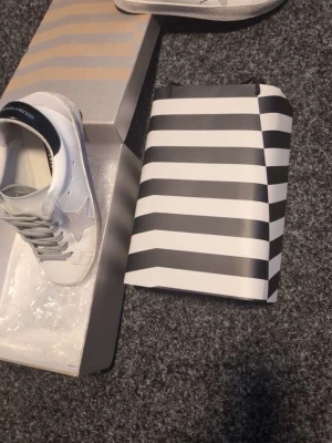 Vita Golden Goose sneakers med stjärna - Golden Goose sneakers i vitt läder med grå mockadetaljer och ikonisk grå stjärna på sidan. Skorna har svarta detaljer vid hälen och grå skosnören. Klassisk låg modell med slitstark sula och ventilerande hål på sidan.