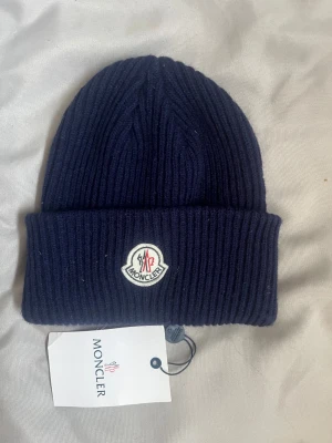 Mörkblå ribbstickad Moncler mössa - Säljer en mörkblå ribbstickad mössa från Moncler med uppvikt kant och klassisk Moncler-logga framtill. Mössan är tillverkad i mjukt material och har en stilren, tidlös design som passar perfekt till kalla dagar.