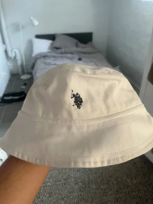 Beige bucket hat från U.S. Polo Assn. - Snygg beige bucket hat från U.S. Polo Assn. med klassisk svart broderad logga framtill. Hatten är tillverkad i bomull och har en enkel, stilren design som passar perfekt till streetwear och sommaroutfits.