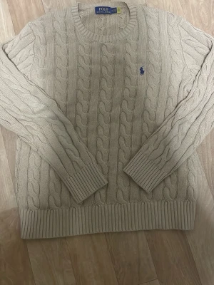 Beige kabelstickad tröja Polo Ralph Lauren - Säljer en beige kabelstickad tröja från Polo Ralph Lauren med rund halsringning och blå broderad logga på bröstet. Tröjan har långa ärmar och ribbade muddar vid ärmslut och nederkant. Klassisk och tidlös design i mjukt material.