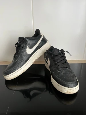 Nike Air Force 1 svarta sneakers - Klassiska Nike Air Force 1 sneakers i svart läder med vit sula och vit swoosh på sidan. Skorna har lågt skaft, svarta skosnören och detaljer i vitt på hälen där det står 'Nike Air'. Perfekta för dig som gillar tidlös streetstil.
