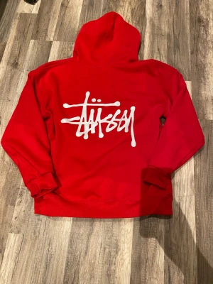 Röd Stüssy hoodie med logga - Snygg röd hoodie från Stüssy med stor vit logga på ryggen och mindre logga på bröstet. Klassisk huva med dragsko och stor magficka framtill. Tillverkad i mjukt material, perfekt för en streetwear-look.