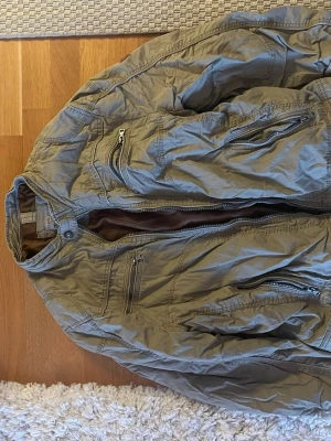 Grön jacka från Angelo Litrico  - Stilren grön vindjacka från Angelo Litrico med flera dragkedjeförsedda fickor framtill. Jackan har hög krage och är tillverkad i ett lätt, vindtätt material som passar perfekt för blåsiga dagar. Snygga sömdetaljer och modern passform.