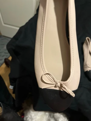 Beige ballerinaskor med svart tå - Säljer ett par snygga ballerinaskor i beige med svart tå och liten rosett framtill. Skorna har rundad tå och klassisk design, perfekta för en stilren look. Materialet ser ut att vara konstläder eller liknande. Passar till många olika outfits!